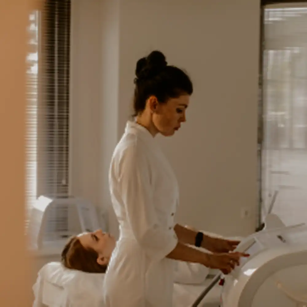 A woman in a white coat operates a medical device while another woman lies on a bed in a clinical room.
